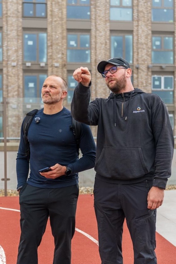 Two men outdoors on a red track, with the man on the right pointing at something ahead while the other watches.